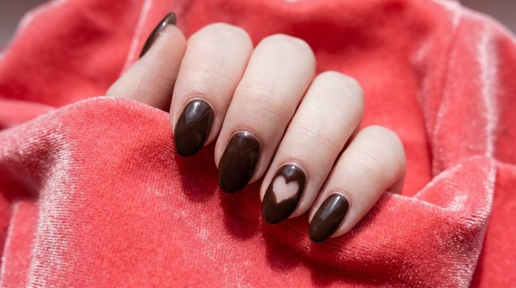 Short almond dark brown nails with a small clear heart cutout accent on an eighteen year old female hand with very white skin over coral pink velvet.