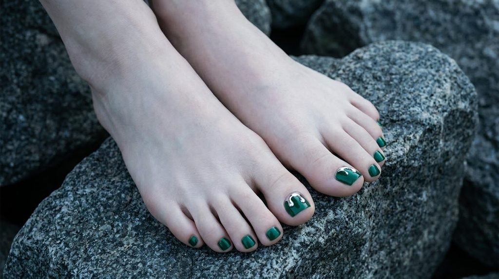 Dark emerald green toenails with silver chrome details on smooth white feet against stone.