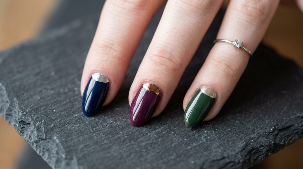 Close-up of three almond nails on one hand, each showing a different color combination: navy/silver chrome, plum/bronze, and forest green/champagne gold in a reverse French style.
