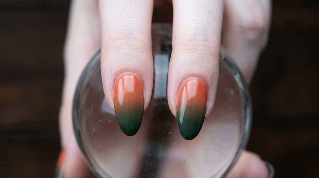 Extreme close-up of two almond nails showcasing a flawless coral-to-deep-hunter-green sunset ombre gradient on a dark brown wood background.