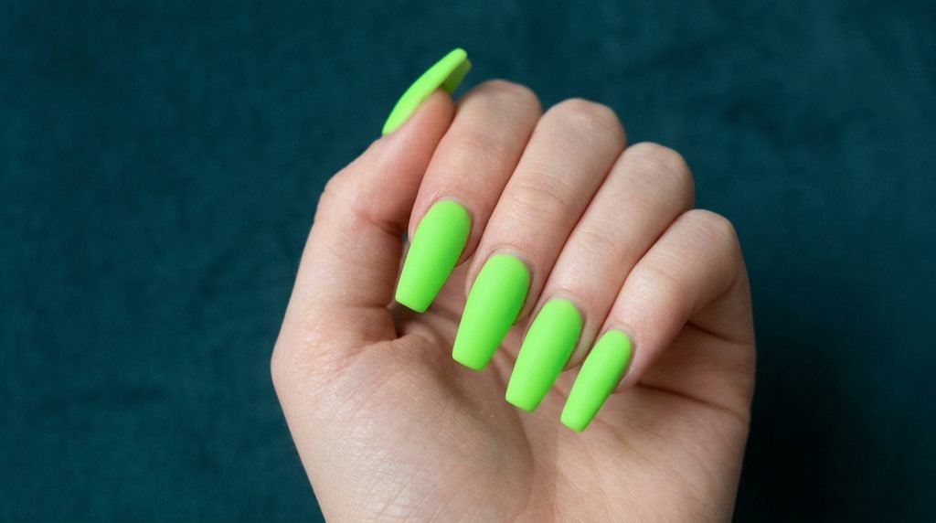 Well-maintained matte neon green coffin nails on an 18-year-old female hand with very white skin over dark teal fabric.