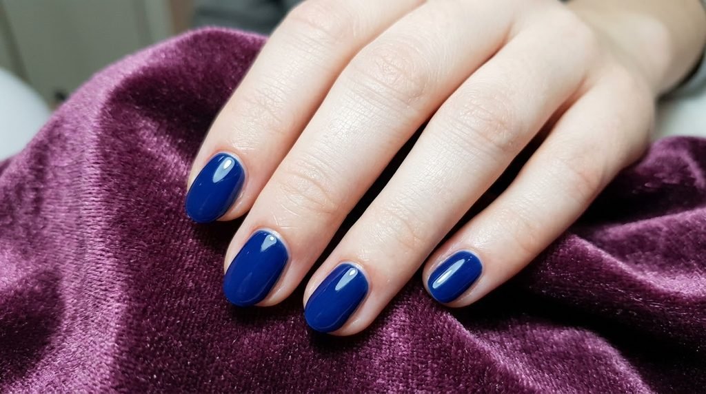 Perfect cuticle application on sapphire blue oval nails on very white female skin over dark plum velvet.