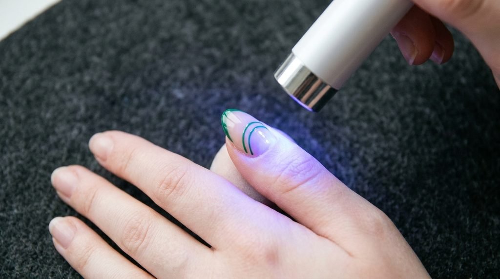 Mini handheld LED light flash curing a freshly painted green double French tip line on a nail.