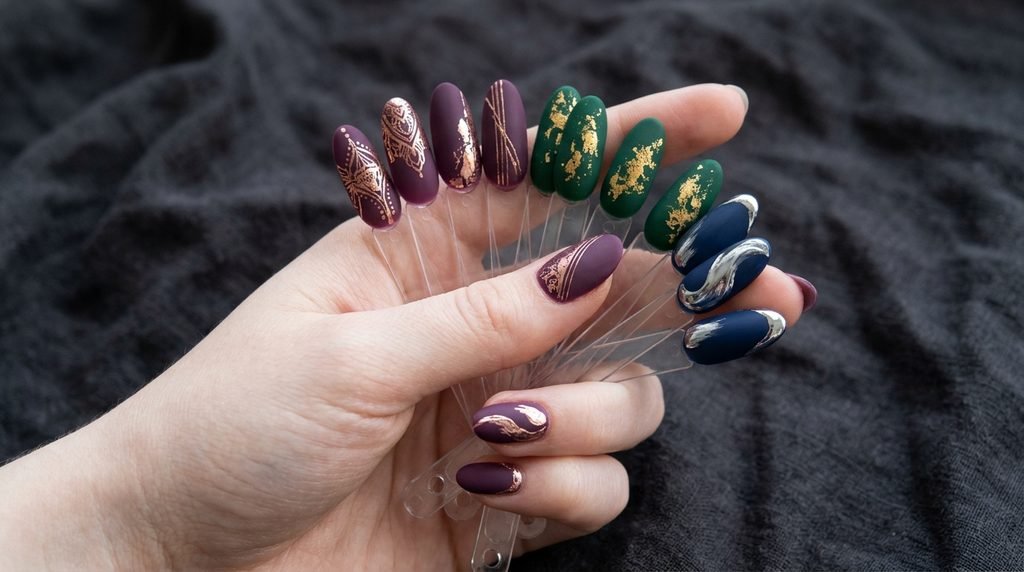 Matte plum, emerald, and navy sample nails with metallic accents held by an 18-year-old female hand with very white skin against dark charcoal.