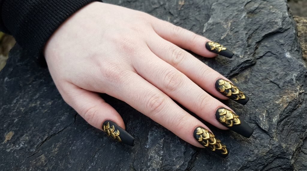Matte black and gold snake scale coffin nails on a pale hand over dark stone background.