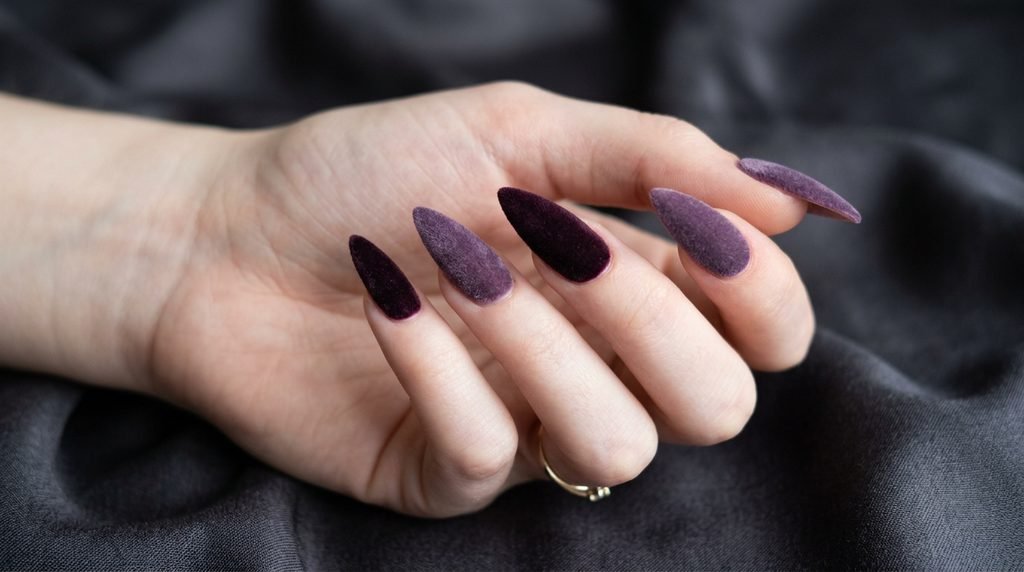 Close-up of stiletto nails on a very white female hand, 18, showing a mix of plum noir and smoky amethyst velvet finishes against charcoal silk.