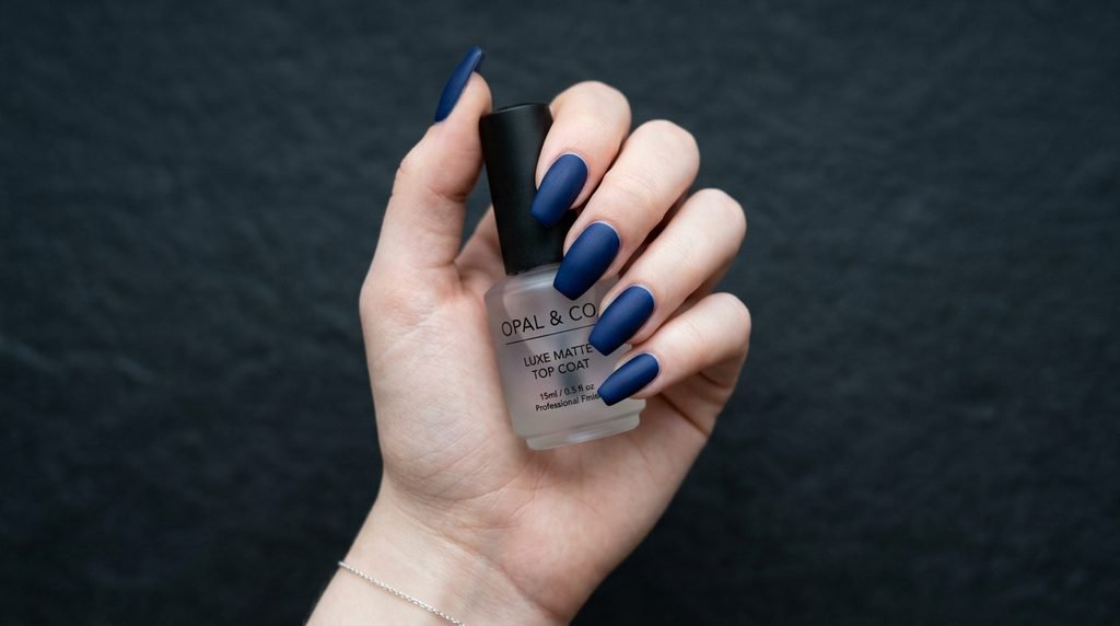 A close-up of a smooth-skinned 18-year-old female hand with very white skin holding a clear top coat bottle, with medium coffin matte navy nails.