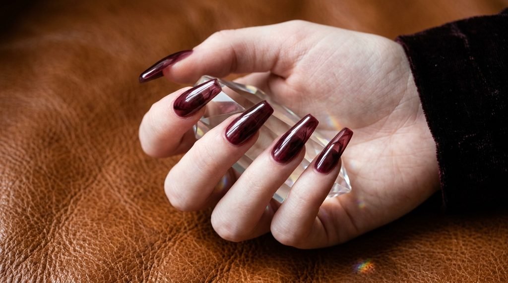 Long coffin extensions with sheer red glass tint on fair hand over brown cognac leather.