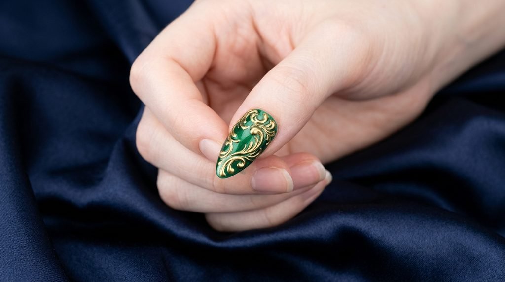 Press-on nail with gold filigree on emerald base held by very white female hand over navy satin.