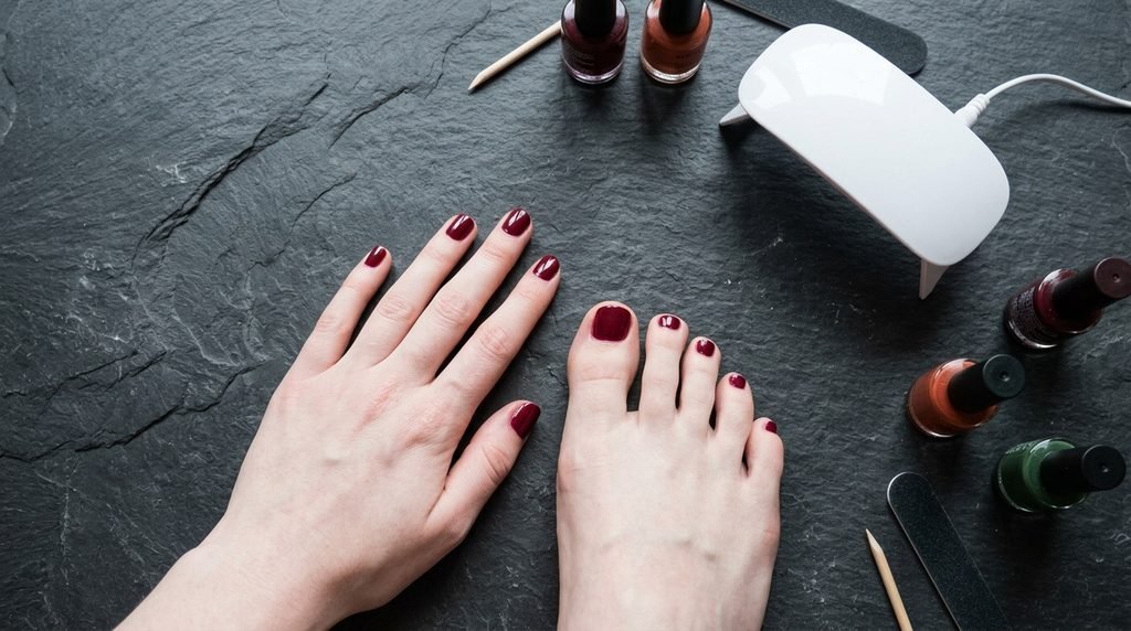 Female hand and foot with white skin next to compact white LED nail lamp and fall polish bottles on slate desk.