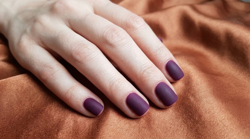 Short square female nails with smooth matte deep plum purple polish resting against a copper satin backdrop.