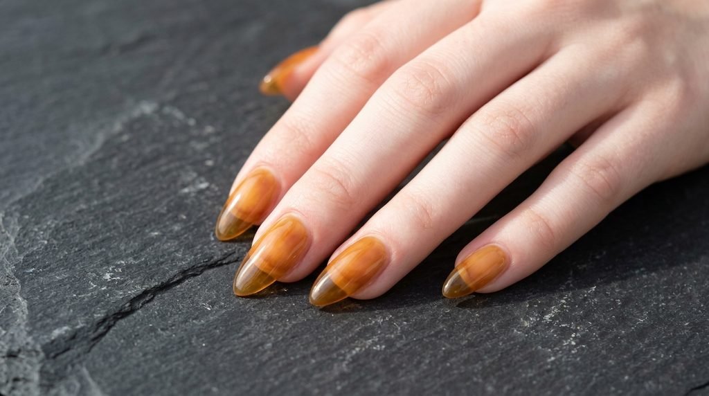 Amber jelly translucent almond nails on an 18-year-old female hand with very white skin tone against a dark charcoal slate surface.