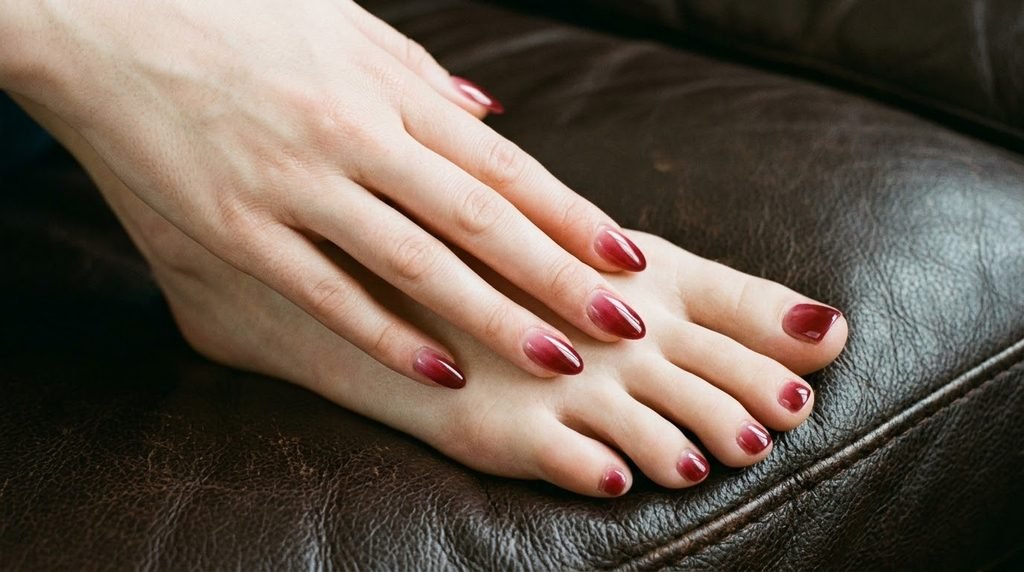 Translucent cranberry syrup ombré almond nails on female hand resting over toes on dark leather.
