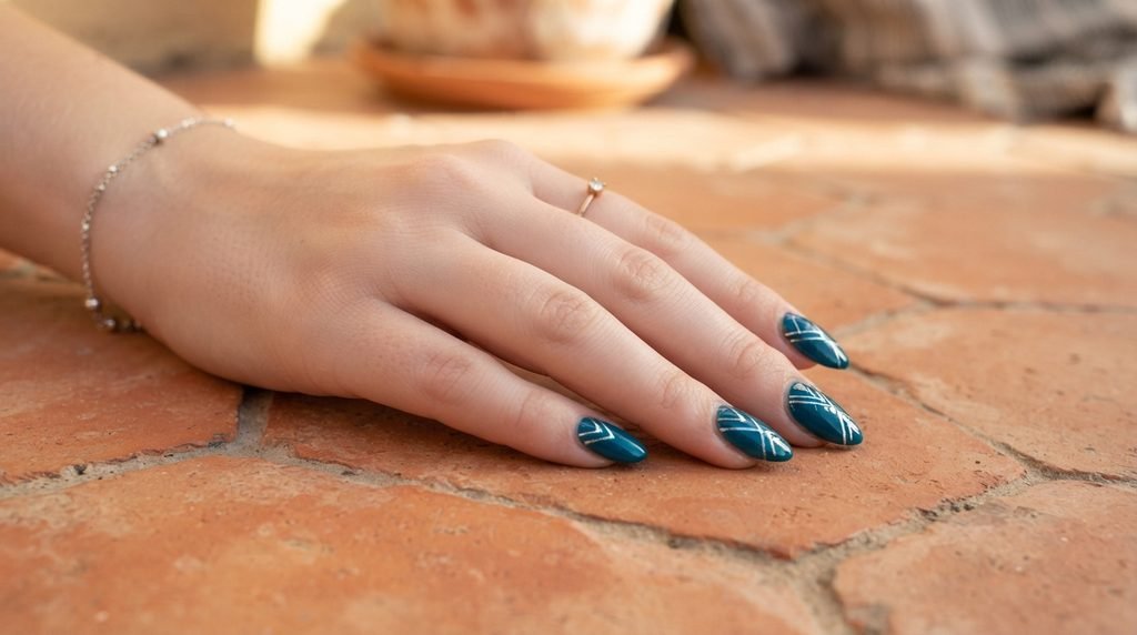 Close-up of an 18-year-old female hand with white skin displaying deep teal almond nails with silver geometric accents on terracotta tile.