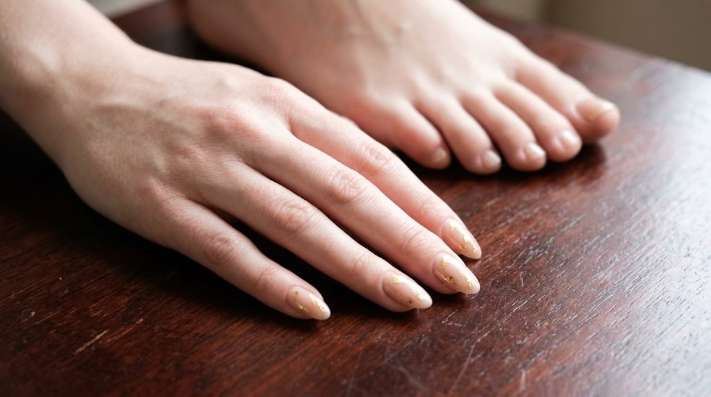 Soft almond nails with neutral polish and gold foil accents on female hand resting near toes on white skin.