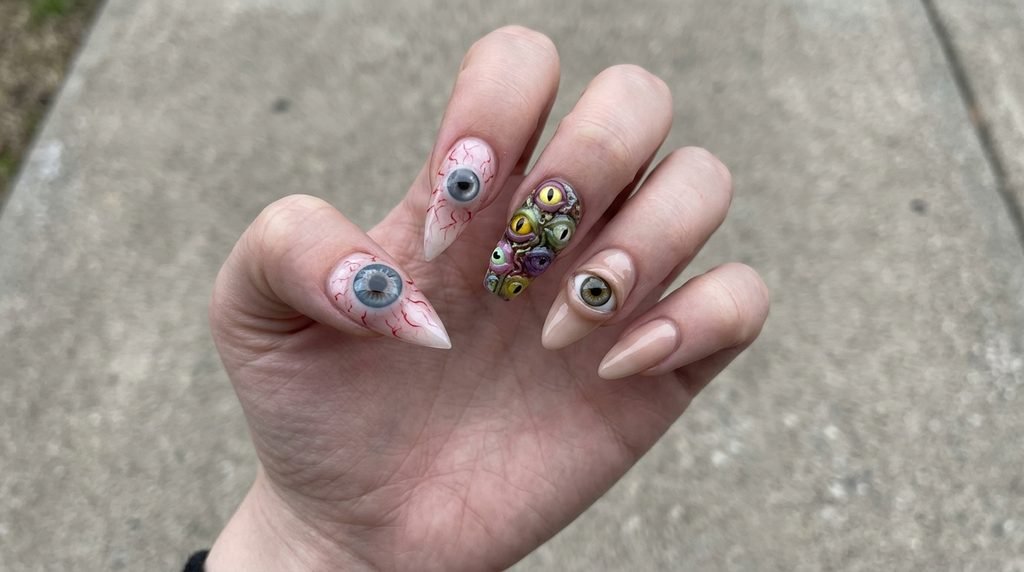 A comparative close-up of a woman's pale hand showing three different nail shapes: a stiletto with a large 3D eyeball, a coffin with multiple eyes, and an almond with a single eye, against blurred concrete.