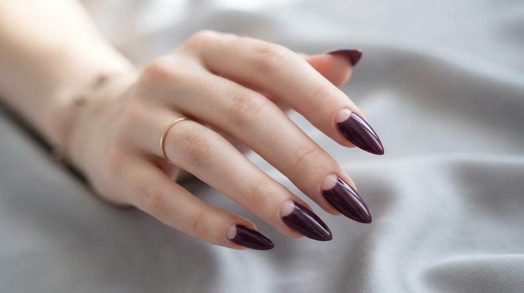 Close-up of plum noir stiletto nails with negative space details on a very white female hand, 18, against blurred light grey silk.