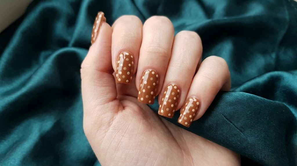 Square caramel nails with cream fawn spots displayed against dark teal satin fabric.