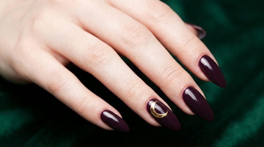 Long almond nails painted deep plum with a delicate gold moon accent on an 18-year-old female hand with white skin.