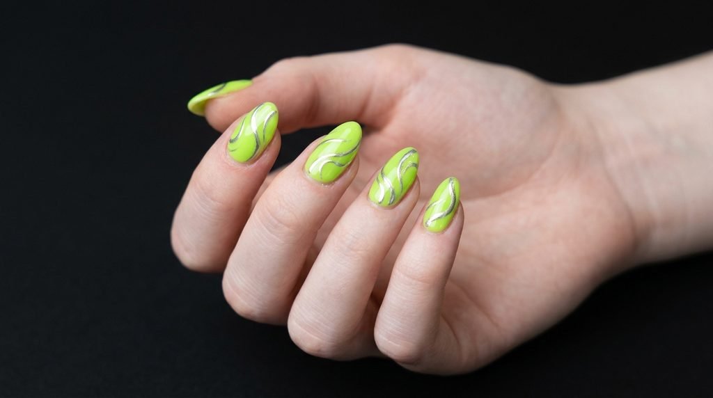 Medium almond nails featuring neon lime green polish and silver metallic swirls on white skin.