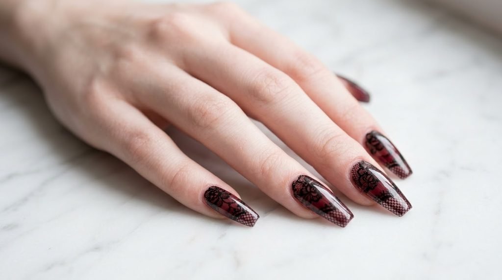 Intricate black lace nail art painted over sheer burgundy coffin nails against a white marble background.