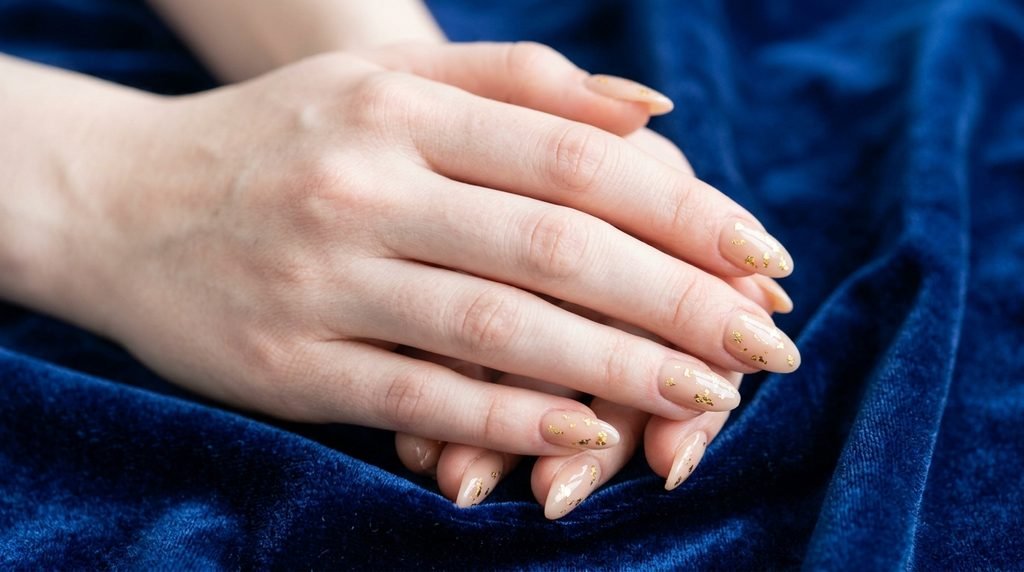 Nude press-on nails with gold foil accents on an 18-year-old female hand with very white skin against sapphire velvet.
