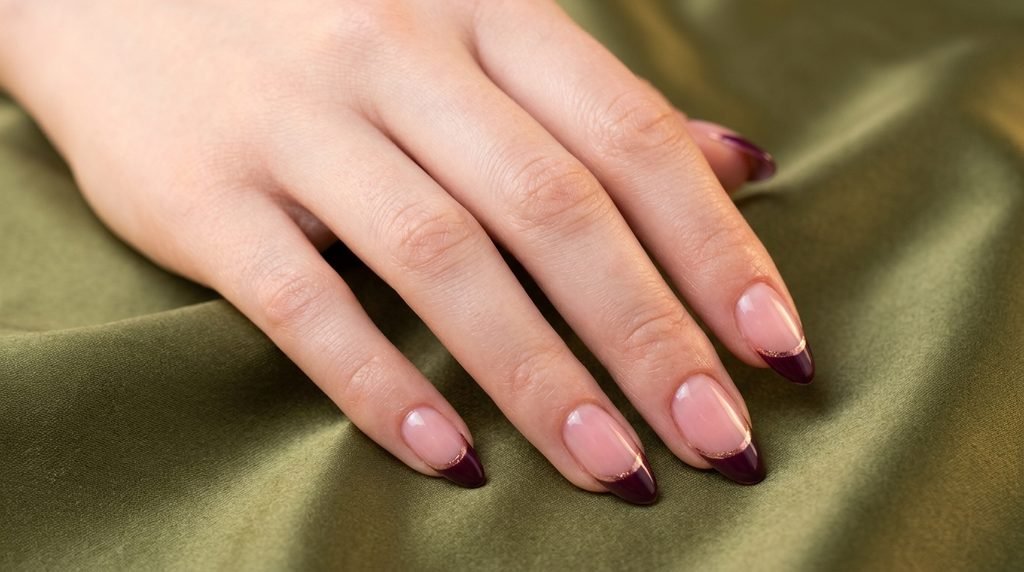 Almond nails with burgundy and rose gold double French tips against an olive green satin backdrop.