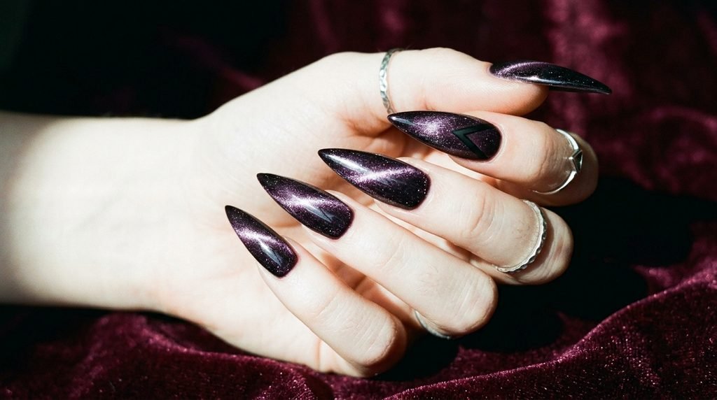 Sharp plum noir velvet stiletto nails with high-contrast magnetic lines on a very white female hand, 18, against deep burgundy velvet.