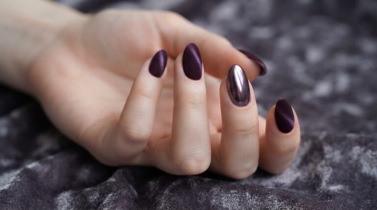 Close-up of almond nails on a very white female hand, 18, showing deep plum velvet and one smoky amethyst chrome accent nail against grey-purple fabric.