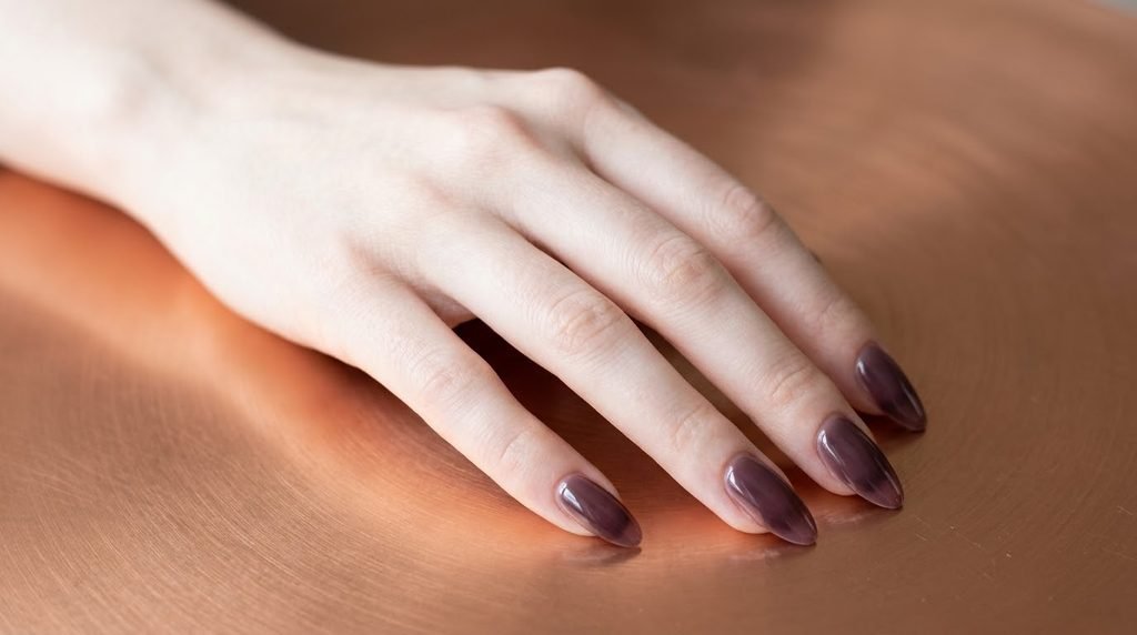 Cool mauve plum syrup nails on fair skin resting over smooth copper plate background.