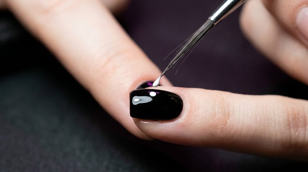 Macro close-up of a very fine-tipped liner nail brush loaded with white paint, poised just above a short square nail of an 18-year-old female hand with smooth white skin, which is painted with black polish, set against a dark charcoal background with violet undertones.