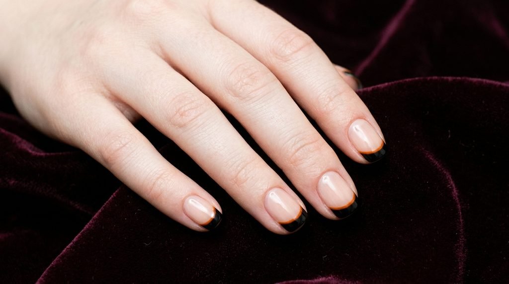 Short squoval nails on a female hand with very white skin displaying two-tone black and  orange French tips.