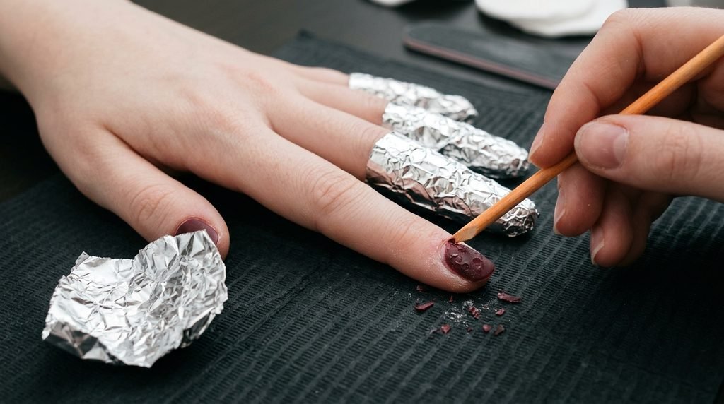 An 18-year-old female hand with very white skin removing softened matte gel with a wooden stick against a dark neutral background.