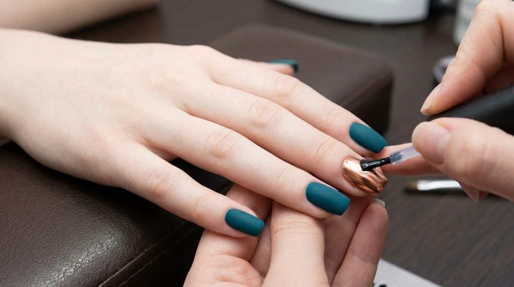 Close-up of a clear top coat brush applying shine over copper chrome on an 18-year-old female hand with very white skin and matte teal nails.