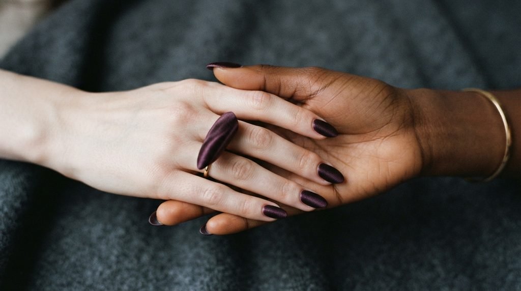 Close-up of two 18-year-old female hands, one very white, one deep-toned, both wearing plum noir velvet almond nails.