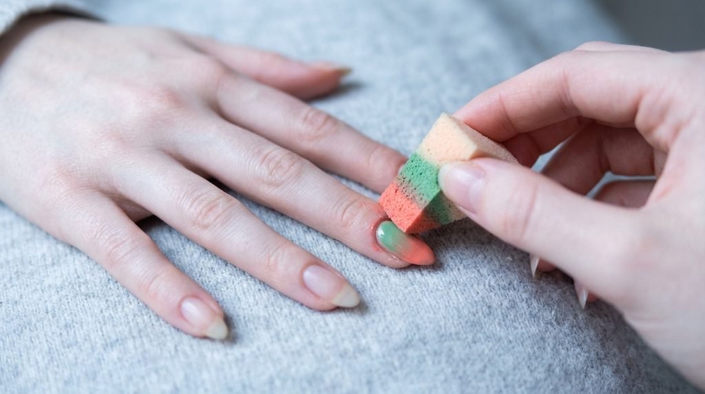A precise close-up of a young woman's hand holding a gel polish brush with minimal product near an almond nail, against a deep emerald green textured background.