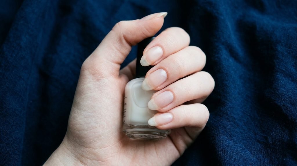 A young woman’s hand with clean, almond nails holding a milky white polish bottle against a deep navy blue background.