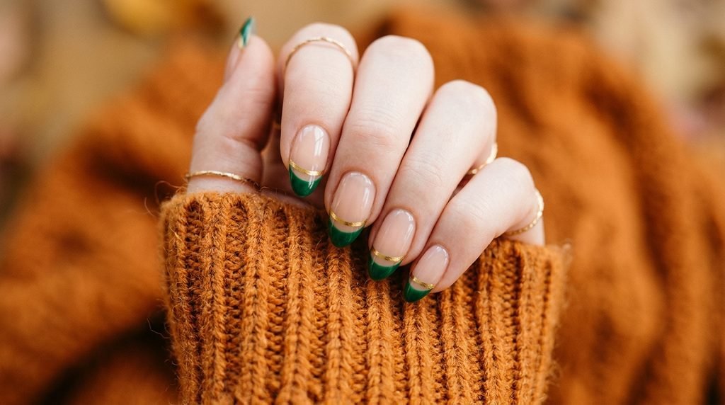 Female hand wearing a burnt orange sweater with emerald and gold double French tip nails.