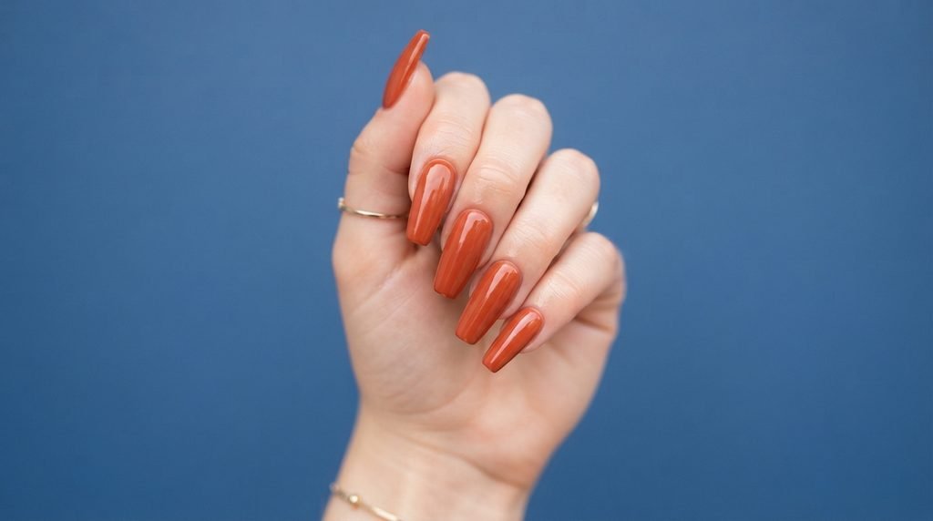 Close-up photograph of high-gloss terracotta cream painted coffin nails on a young woman's hand, fingers vertical, against a matte blue background.
