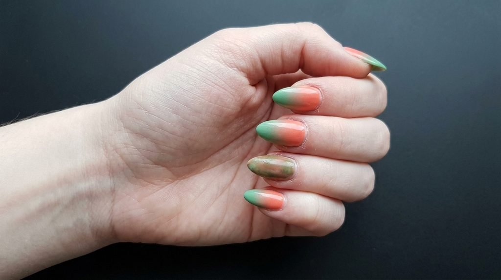 A close-up view of a young woman's hand with almond nails, where the coral-to-green gradient looks muddy on one nail, against a smooth black matte background.