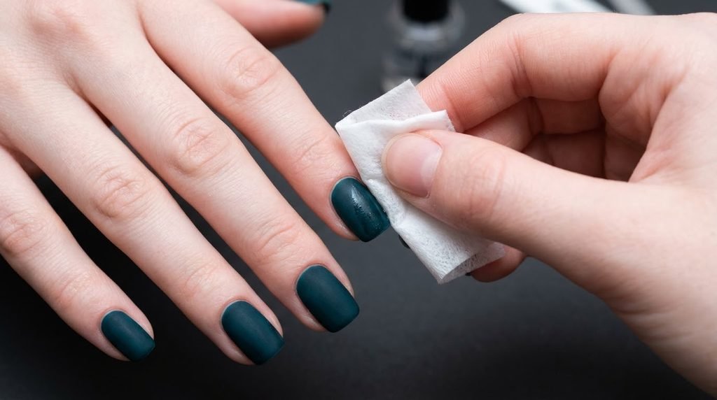 An 18-year-old female hand with very white skin using an alcohol wipe on a matte teal nail against a dark neutral background.