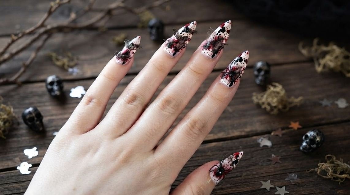 A close-up image of long stiletto nails on an 18-year-old female hand with white skin, featuring a crisp white base covered in random red blood splatter droplets. One nail has a thick drip. Hand holds a small skull against dark velvet. High-definition.