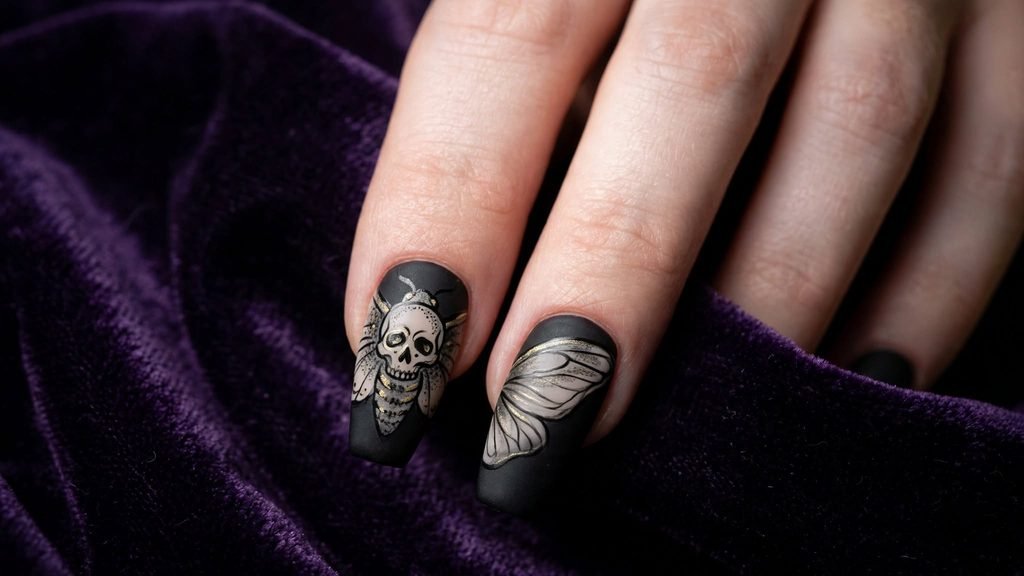 Close-up of two matte black coffin nails featuring an intricately painted skull detail inspired by a moth's thorax, with gold and grey dotting. Posed on dark purple fabric with dramatic side lighting.