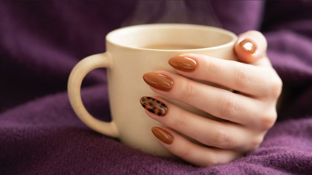 Close-up of glossy caramel toffee almond nails with a tortoise shell accent nail on a young woman's hand against deep purple cashmere.